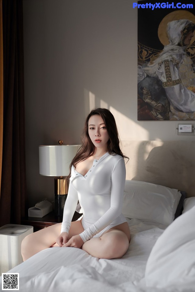 A woman sitting on a bed in a white bodysuit.